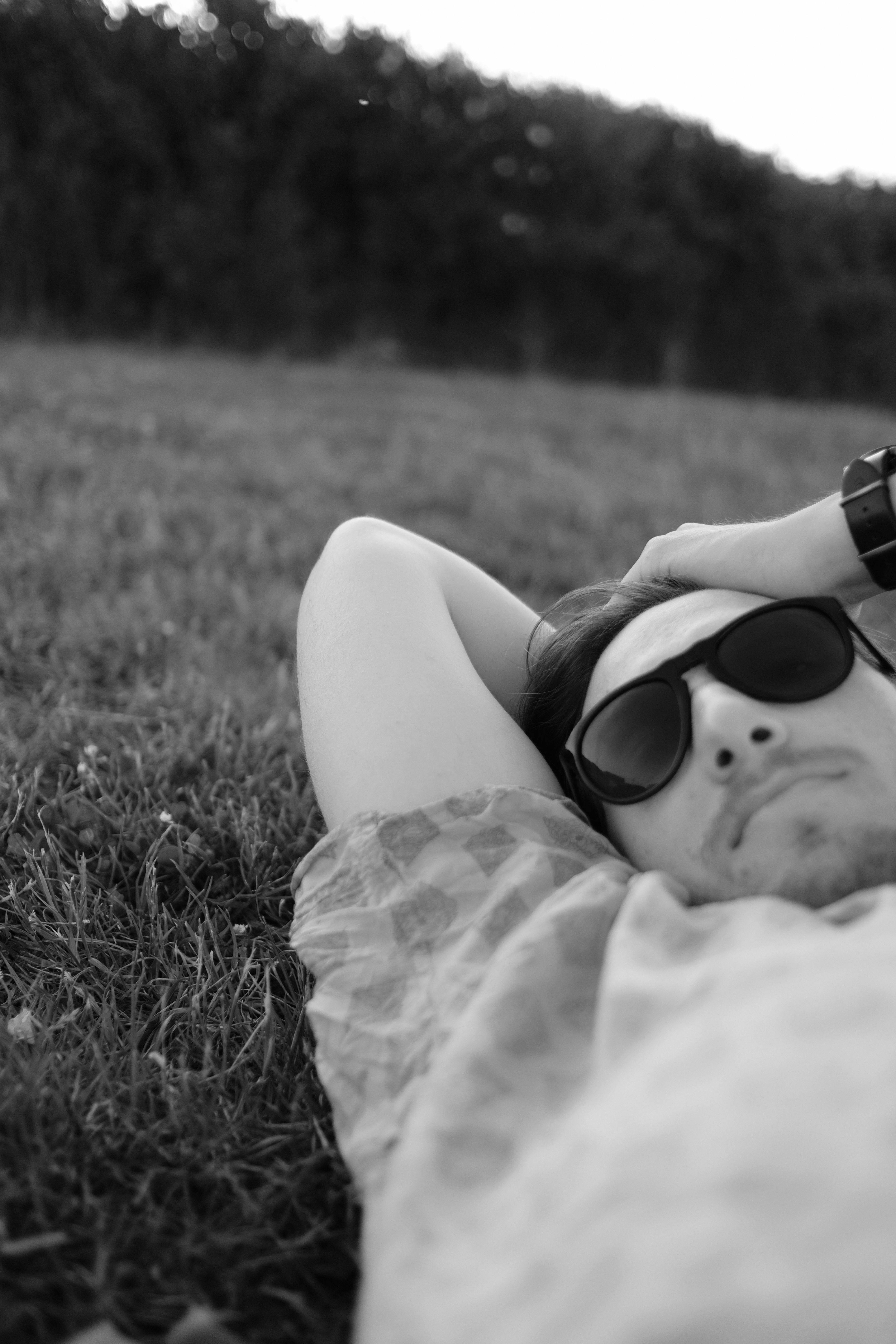 A man in sunglasses lays in the grass, holding his arm behind his head looking towards the camera.